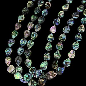 May include: A collection of teardrop-shaped abalone shell beads arranged in multiple strands. Each bead displays a unique iridescent surface with swirling patterns of deep green, blue, purple, and silver hues against a solid black background.