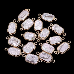 May include: A collection of rectangular, white, baroque pearl pendants with gold-colored metal edging and small loops for stringing. The pearls have an irregular, organic shape and a lustrous surface. The background is black.