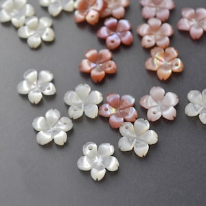 May include: A collection of flower-shaped beads in various shades of white, pink, and peach. The beads have five petals and a small hole in the center, suggesting they are for jewelry making or crafting. The beads are scattered on a gray surface.