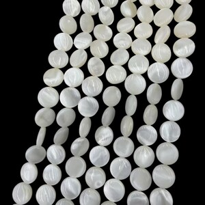May include: A collection of white mother of pearl beads arranged in multiple vertical strands. Each bead is circular, flat, and polished, displaying a natural iridescent sheen and smooth surface texture against a solid black background.
