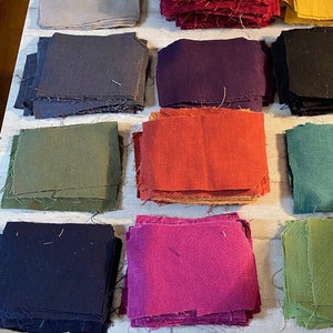 Peut inclure: Des piles de carrés de tissu de différentes couleurs, dont le rouge, l'orange, le jaune, le vert, le bleu, le violet et le noir. Les carrés de tissu sont disposés en rangées sur une surface blanche. Le chiffre "0" est écrit en jaune sur l'un des carrés de tissu.