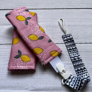 May include: Two pink fabric burp cloths with a lemon print and a white clip. A black and white geometric patterned pacifier clip is attached to the burp cloths.