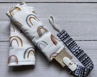 May include: Two padded fabric covers featuring a white background with repeating rainbow arches in shades of brown, tan, and gray, accompanied by a matching fabric strap with a black and white geometric pattern and a white plastic clip.