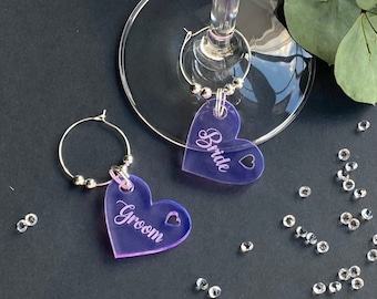 May include: Two purple heart-shaped acrylic wine glass charms with the words "Bride" and "Groom" engraved on them. The charms are attached to silver hoops with silver beads.