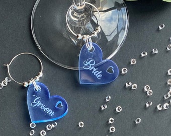 May include: Two blue heart-shaped acrylic wine glass charms with silver rings. The charms are engraved with the words "Bride" and "Groom".