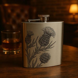 May include: A silver-coloured hip flask featuring a black thistle illustration. The flask has a hinged cap and a brushed metal finish. A glass of amber liquid and a dark wooden surface are in the background.