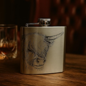 May include: A stainless steel hip flask featuring an etched Highland cow design. The flask has a curved shape and a silver cap. The background is a blurred wooden surface.