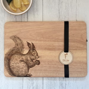 Op de afbeelding: Een houten snijplank met een lasergegraveerd beeld van een eekhoorn die een noot vasthoudt. De plank heeft een zwart lint eromheen gebonden met een rond label waarop "Elle & Hart" staat.