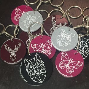 May include: A set of 10 keychains with different geometric animal designs. The keychains are made of acrylic and have a burgundy, gray, or black background. The designs include a deer, butterfly, wolf, and fox. The keychains are attached to silver key rings.