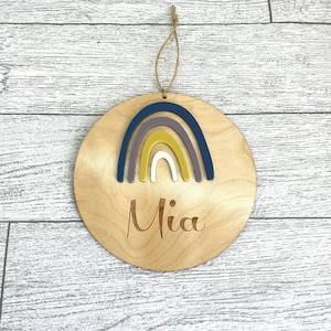 Puede incluir: Un letrero redondo de madera con un arcoíris hecho de fieltro superpuesto en tonos de azul, morado, amarillo y blanco. El nombre "Mia" está grabado en el letrero.