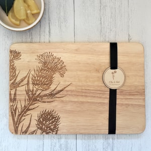 May include: A wooden cutting board with a laser-engraved design of thistles. The board has a black ribbon tied around it with a round wooden tag that says "Ella & Mae".
