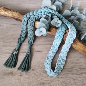 Op de afbeelding: Een gevlochten lichtblauw koordketting met twee bungelende kwastjes aan de uiteinden, elk versierd met een kleine zilverkleurige ronde bedel die een boom voorstelt. Het koord is gemaakt van een dik, geweven materiaal met een textuur.