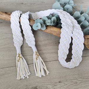 Op de afbeelding: Een lange, gevlochten riem gemaakt van wit en cr&egrave;mekleurig koord. De riem heeft twee uiteinden afgewerkt met decoratieve goudkleurige metalen bedels en bungelende franjes, wat zorgt voor een getextureerd, geweven uiterlijk over de gehele lengte.