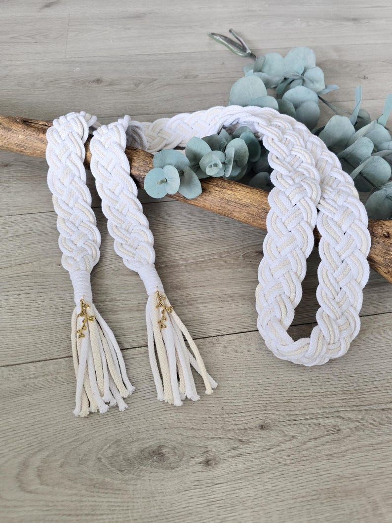 Op de afbeelding: Een lange, gevlochten riem gemaakt van wit en cr&egrave;mekleurig koord. De riem heeft twee uiteinden afgewerkt met decoratieve goudkleurige metalen bedels en bungelende franjes, wat zorgt voor een getextureerd, geweven uiterlijk over de gehele lengte.