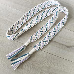 Op de afbeelding: Een lange, gevlochten koordriem met een geweven patroon in cr&egrave;me, lavendel en groenblauw. De riem eindigt in twee bijpassende kwastjes die zijn vastgezet met zilverkleurige metalen accenten, wat een decoratief accessoire met een textuurontwerp vormt.