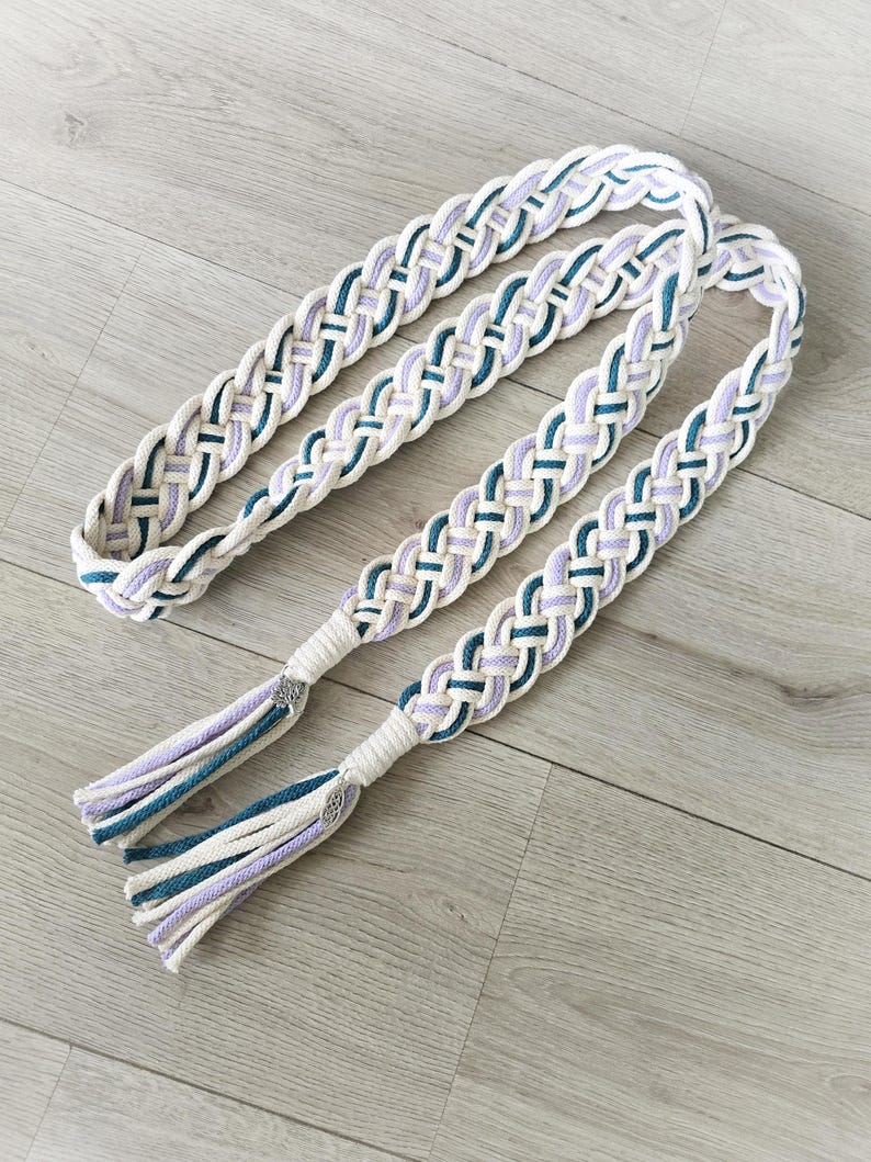 Op de afbeelding: Een lange, gevlochten koordriem met een geweven patroon in cr&egrave;me, lavendel en groenblauw. De riem eindigt in twee bijpassende kwastjes die zijn vastgezet met zilverkleurige metalen accenten, wat een decoratief accessoire met een textuurontwerp vormt.