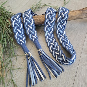 Op de afbeelding: Een gevlochten koordriem met een geweven patroon van donkerblauwe, lichtblauwe en witte strengen. De riem eindigt in twee kwastjes, elk bevestigd met een goudkleurige halve maan bedel, wat een decoratief accessoire met een meerkleurig ontwerp vormt.