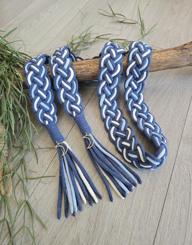 Op de afbeelding: Een gevlochten koordriem met een geweven patroon van donkerblauwe, lichtblauwe en witte strengen. De riem eindigt in twee kwastjes, elk bevestigd met een goudkleurige halve maan bedel, wat een decoratief accessoire met een meerkleurig ontwerp vormt.