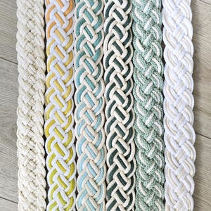 Op de afbeelding: Zes lange, platte, gevlochten textielstroken verticaal gerangschikt. De kleuren omvatten effen cr&egrave;me, geel en wit, lichtblauw en wit, donkergroen en wit, saliegroen en wit, en een tweekleurig cr&egrave;me en wit gevlochten patroon.