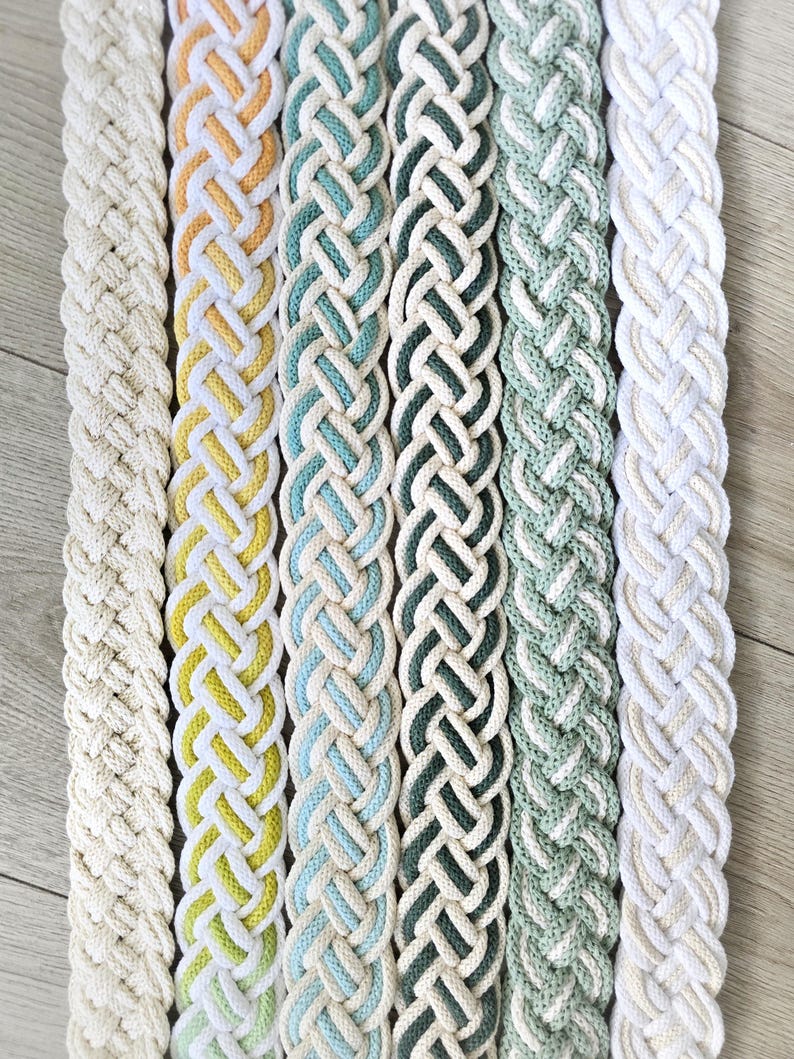 Op de afbeelding: Zes lange, platte, gevlochten textielstroken verticaal gerangschikt. De kleuren omvatten effen cr&egrave;me, geel en wit, lichtblauw en wit, donkergroen en wit, saliegroen en wit, en een tweekleurig cr&egrave;me en wit gevlochten patroon.