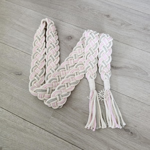 Op de afbeelding: Een gevlochten riem met een geweven patroon van lichtroze, zachtgrijs en gebroken wit koord. De riem eindigt in twee lange kwastjes, met een kleine zilverkleurige zonvormige bedel bevestigd aan de basis van een kwastje om de strengen vast te zetten.