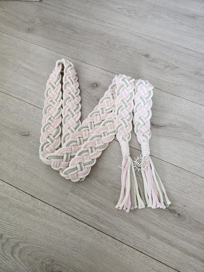 Op de afbeelding: Een gevlochten riem met een geweven patroon van lichtroze, zachtgrijs en gebroken wit koord. De riem eindigt in twee lange kwastjes, met een kleine zilverkleurige zonvormige bedel bevestigd aan de basis van een kwastje om de strengen vast te zetten.