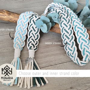 Op de afbeelding: Gevlochten koordriemen met witte, lichtblauwe en donkergroene strengen met kwastjes. E&eacute;n riem heeft een zilverkleurige hartvormige bedel. Het logo toont MIDGARA CORDS & BELTS en tekstlabels voor de binnen- en buitenste strengen.