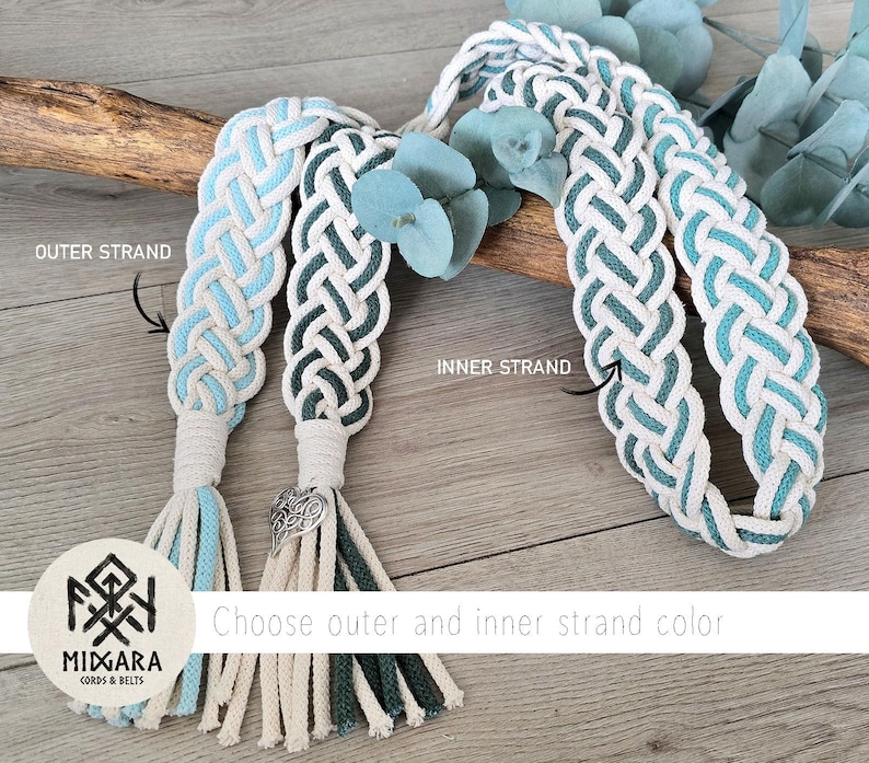 Op de afbeelding: Gevlochten koordriemen met witte, lichtblauwe en donkergroene strengen met kwastjes. E&eacute;n riem heeft een zilverkleurige hartvormige bedel. Het logo toont MIDGARA CORDS & BELTS en tekstlabels voor de binnen- en buitenste strengen.