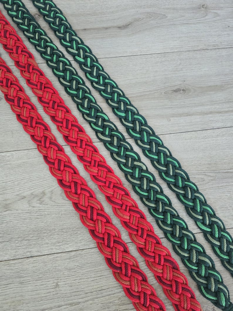 Op de afbeelding: Twee lange, platte, gevlochten textielbanden die diagonaal zijn gerangschikt. De ene band heeft een verloop van rode, oranje en diepe bordeauxrode tinten, terwijl de andere band een verloop van donker bosgroen, smaragd en licht limoengroen toont.