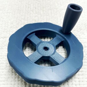 May include: A navy blue, 3D-printed handwheel with a cross-shaped center and a handle. The wheel has a scalloped edge and a central hole. The handle is cylindrical and angled. The object is likely a component for a machine or device.