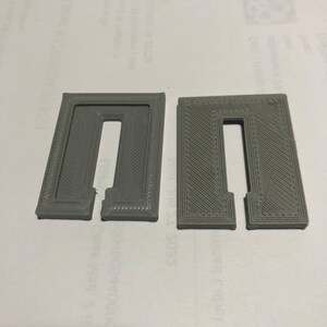 May include: Two gray plastic 3D printed objects with a rectangular shape and a cut-out in the center. The objects have a textured surface with a diagonal pattern.