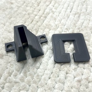 May include: Two 3D printed plastic pieces in gray. One piece is a rectangular shape with a cut-out in the center. The other piece is a triangular shape with a slot cut into the top.