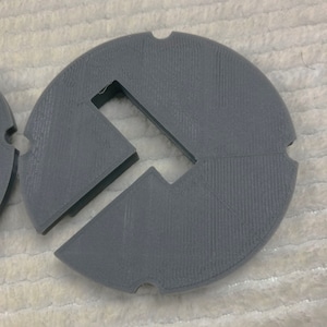 May include: A gray plastic 3D printed circle with a rectangular cutout in the center. The circle has a textured surface.