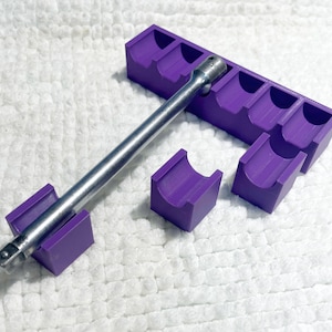 May include: A purple socket organiser with multiple slots, holding a silver metal wrench. Two additional purple holders are also present. The items are arranged on a white, textured surface.