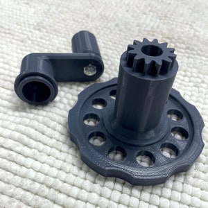 May include: A grey plastic gear with a handle. The gear has a circular base with multiple holes and a smaller gear on top. The handle is a curved piece of plastic with a hole in the end.