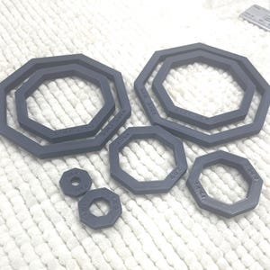 May include: A collection of gray, octagonal rubber gaskets of varying sizes. Some gaskets are nested within each other. The gaskets have printed text indicating size and specifications. The items are arranged on a white, textured surface.
