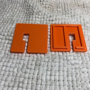 May include: Two orange plastic pieces with a slot cut out of them. The pieces are designed to fit together to create a small box.