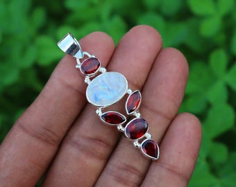 May include: A silver pendant with a large oval moonstone and smaller red garnet stones. The pendant is on a silver chain.