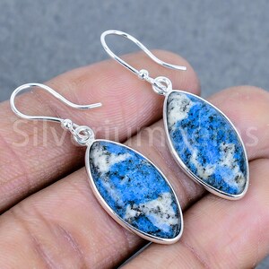 Può includere: Orecchini in argento con pendenti ovali in pietra k2 blu. La pietra presenta un mix di motivi blu, bianchi e neri. Gli orecchini hanno un semplice design a gancio. La parola "Silverium" è parzialmente visibile.