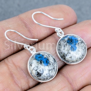Peut inclure: Une paire de boucles d'oreilles pendantes en argent. Chaque boucle d'oreille présente une pierre ronde et polie avec un motif moucheté blanc et gris, rehaussé de taches bleues vives. Les boucles d'oreilles ont un design simple en forme de crochet.