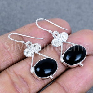 May include: A pair of silver earrings with oval, black gemstones. The earrings have a hook design with a decorative swirl at the top. The gemstones are set in a silver bezel.