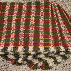 May include: A stack of woven napkins with a red, green, and white checkered pattern. The napkins have a fringe border in green, red, and yellow. The napkins are folded and stacked on top of each other.