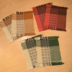 May include: Four woven coasters in shades of brown, orange, and green. Each coaster features a patchwork design with fringe detailing. The coasters are square and appear to be handmade, showcasing a rustic aesthetic.