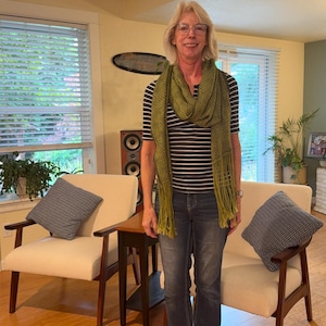 May include: A long, olive green knit scarf featuring a textured, woven pattern and long, straight fringe at both ends. The scarf is draped loosely around the neck, hanging down the front of a dark blue and white horizontal striped short-sleeve shirt.