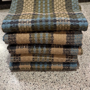 May include: A stack of five woven kitchen towels. The towels have a repeating pattern of squares and rectangles in shades of blue, green, and tan. The towels are neatly folded and stacked on top of each other.