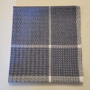 May include: A white cotton tea towel with a blue geometric pattern. The towel is folded in half and has a white border.