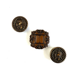 May include: Three decorative buttons with a gold and brown finish. The buttons feature intricate designs with a square and two round shapes.