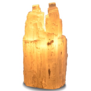 May include: A selenite lamp with a natural, raw, and textured surface. The lamp is lit and emits a warm, soft glow. The selenite is a white, translucent crystal.