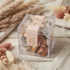 May include: A clear, square gift box filled with foil-wrapped chocolates. A pink band with a gold butterfly design and the words "Mis Quince" wraps around the box. The box is on a light wooden surface with dried flowers.
