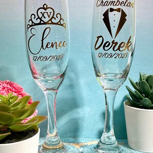 May include: Two clear champagne flutes with gold lettering. One flute has a crown design with the name "Elenee" and the date "12/09/2023". The other flute has a tuxedo design with the name "Derek" and the date "12/09/2023".